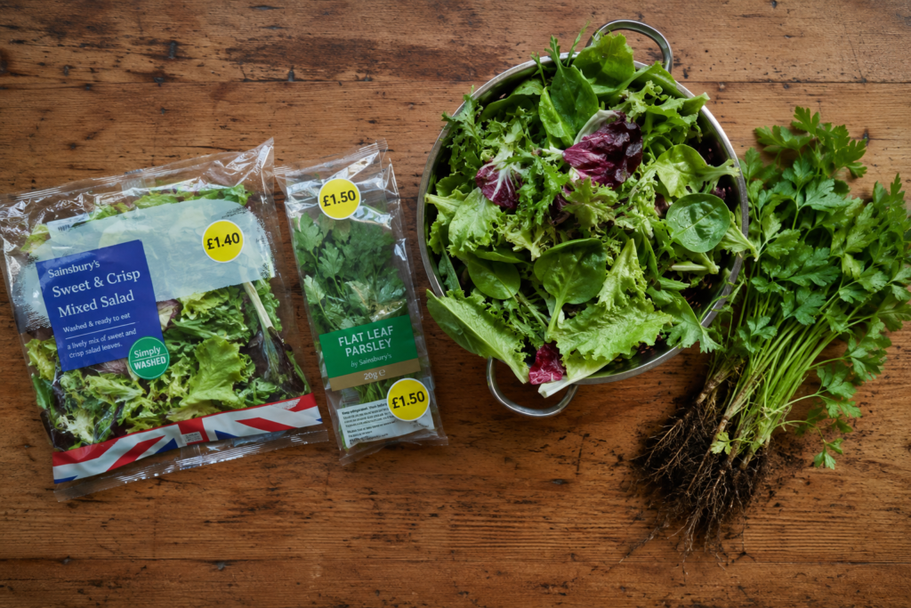 Comparison of supermarket bagged salad and herbs next to a large colander of freshly picked homegrown salad leaves and parsley.